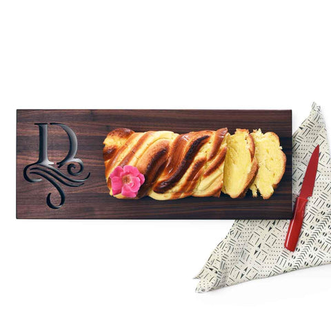 Walnut Wood Long Horizontal Cutting Board with cake-like bread on top. The letter D is cutout in scroll font on the left hand side.