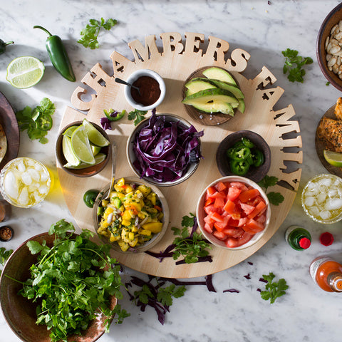 Round Wooden Personalized Cutting Board with names Chamber & Family Cutout on the top. Board is full of food - little bowls of tomatoes and corn salsa, limes and cabbage. Tiny plate with jalapenos, small dish of sauce with spoon and tiny place with sliced avocado.