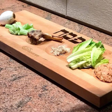 Photograph of Medium Wood Customized Rectangle Seder Plate with engraved family initials in Hebrew at the top - the board is showing only some of the sections - one with celery, one with a shank bone, one with horseradish, one with some lettuce, and one with haroseth.