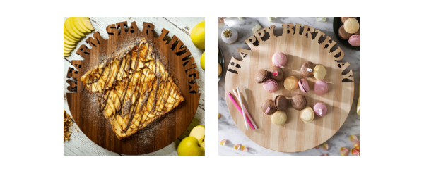 of a rooster and the words Farm Star Living and a star at the end. On top is an apple chocolate tart. The board is setting on a marble surface surrounded by apples.  Next to it is a Round Maple Cutting Board with the words Happy Birthday cut out around the top. On it are chocolate, vanilla, and strawberry macarons and 3 birthday candles.